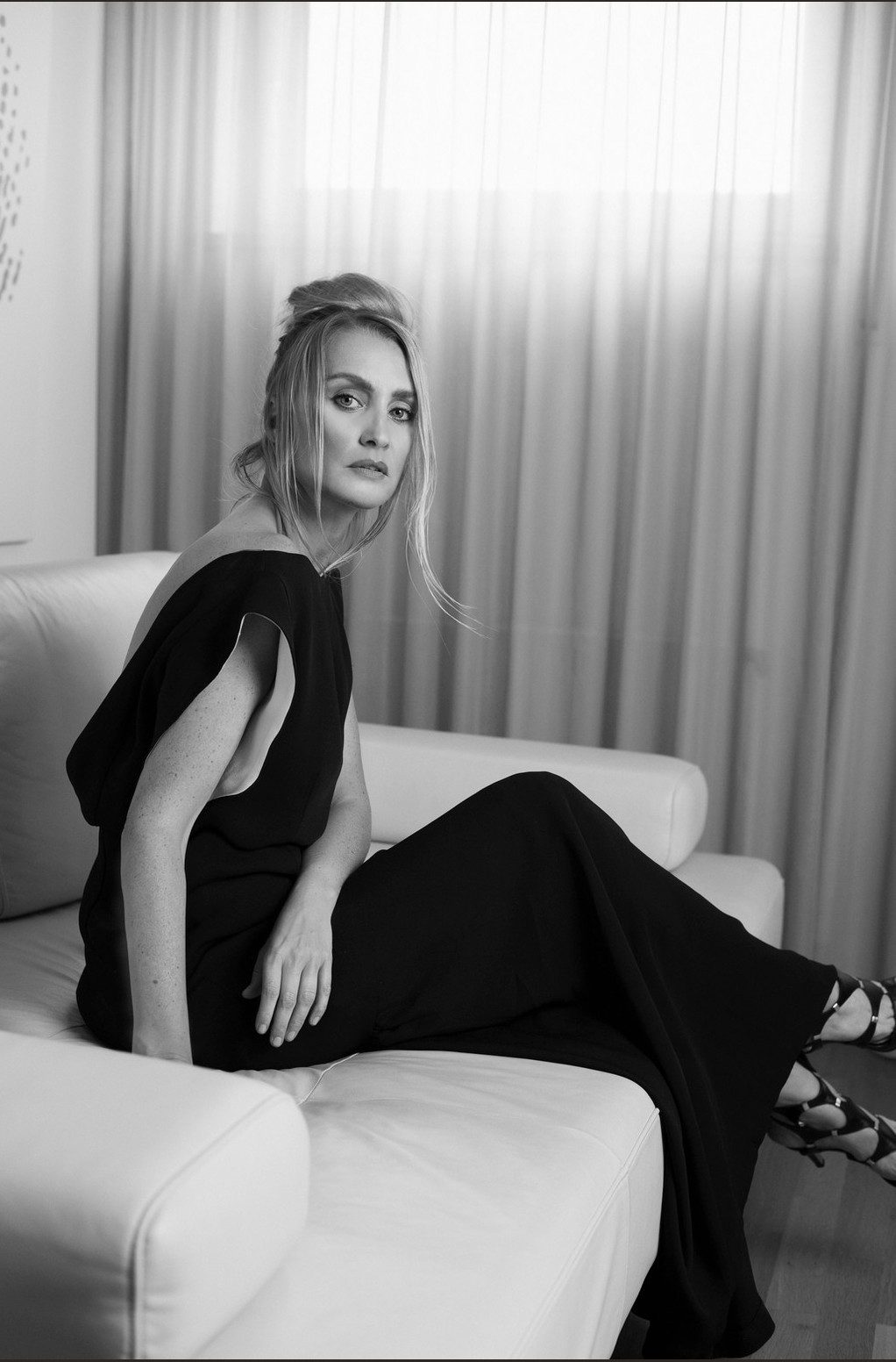 Model im langen schwarzen Kleid auf hellem Sofa vor lichtdurchflutetem Vorhang in Schwarzweiss — Das Studiozwei, Werbefotograf München