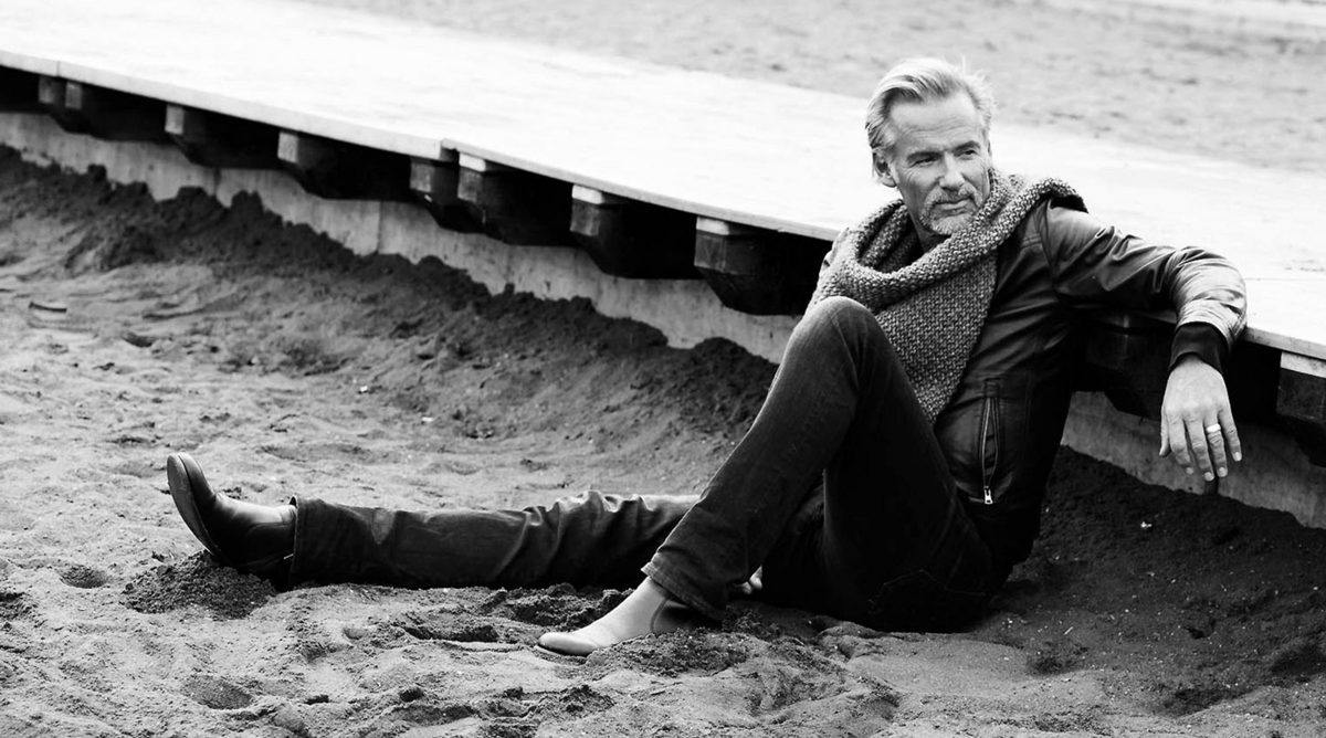 Mann in Lederjacke und Schal sitzt im Sand vor einer Holzplattform, schwarzweiss — Das Studiozwei, Werbefotograf München