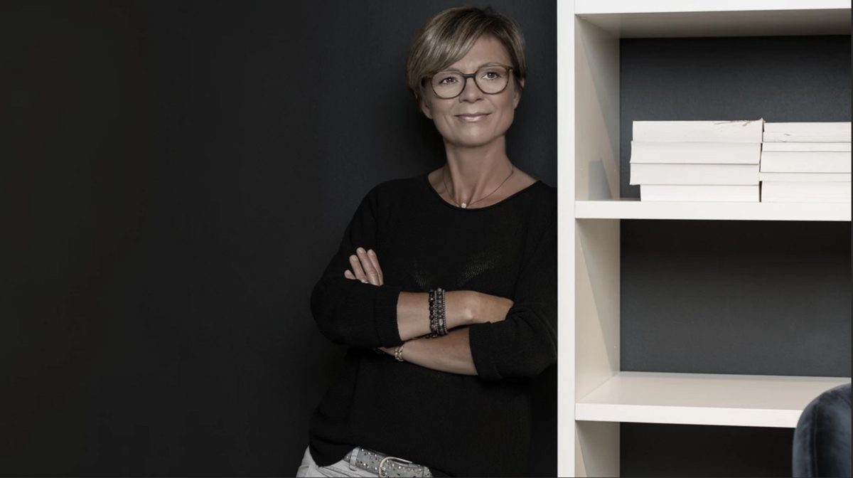Frau mit Brille und verschraenkten Armen lehnt neben einem weissen Buecherregal an dunkler Wand — Das Studiozwei, Werbefotograf München